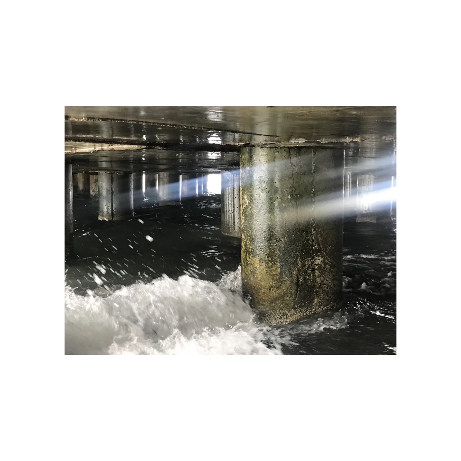 Photograph beneath a pier, waves breaking against mossy concrete pilings crossed by a beam of light
