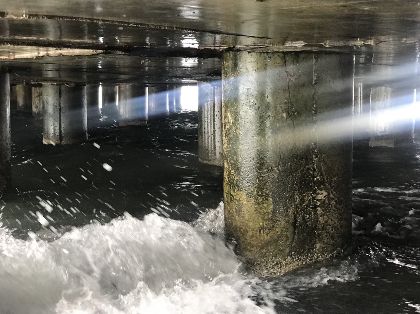 Photograph beneath a pier, waves breaking against mossy concrete pilings crossed by a beam of light
